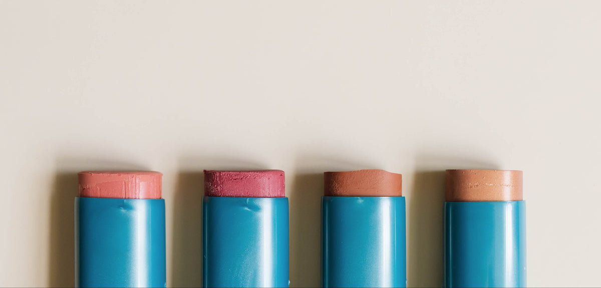 Four blue cosmetic sticks with pink and peach tips arranged vertically on the right side of a light beige background, leaving negative space on the left.