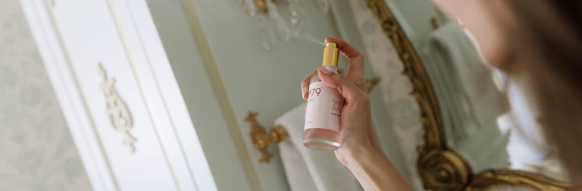 A woman in a bathroom holds a pink spray bottle with a gold cap, spraying it while looking into an ornate gold-framed mirror.